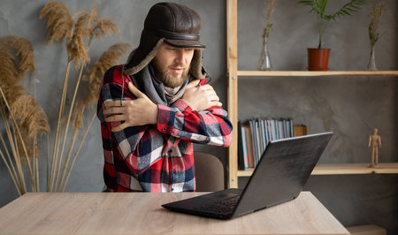 In winter, in a cold apartment without heating. A young Caucasian man wearing a hat and a scarf is working on a laptop at home in a cold apartment.の写真素材