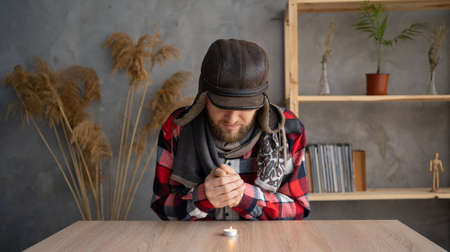 A Caucasian bearded man dressed in a warm winter hat and a scarf sits at home at a table and warms his hands from a burning candle. Concept of no heating in winter at home.の写真素材