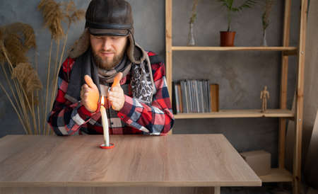 A Caucasian bearded man dressed in a warm winter hat and a scarf sits at home at a table and warms his hands from a burning candle.の写真素材