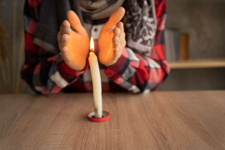 Close-up of male hands warming up near a burning candle which stands on the table. concept of problems with heating, cold at home in winter.の写真素材