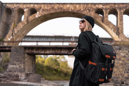 Girl tourist traveler with a backpack standing against the backdrop of an arched bridge, an old railway bridge over the river.の写真素材