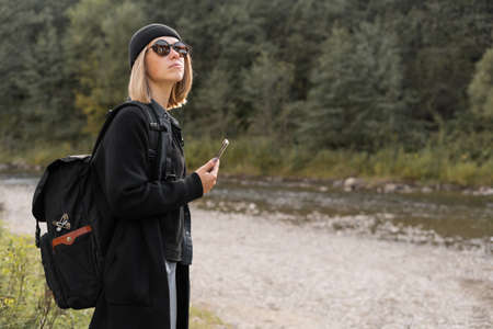 Caucasian girl tourist traveling near the river with a backpack, nature background, stylish young woman traveler in a black coat, side view.の写真素材