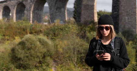 Tourist girl near the old arch bridge, searching for a track online in the phone while traveling, historical architectural structure bridge with a railway.の写真素材