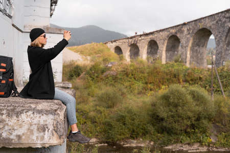 Tourism and travel concept. Girl blogger live stream of the old stone arch bridge, tourist photographing the bridge on the phone.の写真素材