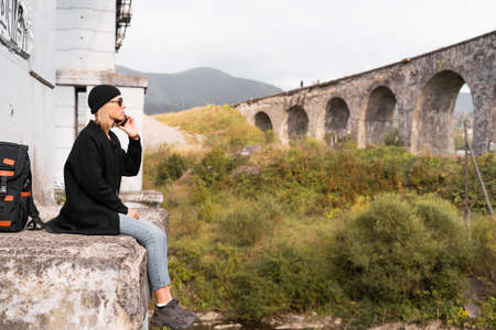 Girl traveler sitting in front of the old stone arch bridge, tourist talking on the phone 5G communication, travel vacation.の写真素材
