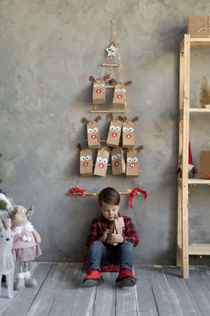 Childrens handmade advent calendar craft bags hanging on the wall, christmas children fan, boy sitting with a gift on the floor, gray wall backgroundの写真素材