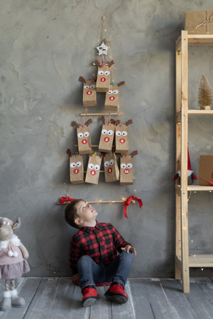 Handmade advent calendar in craft bag hanging on the wall, boy sitting on the floor looking at Christmas presents, horizontal position.の写真素材
