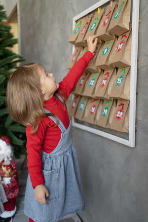 Handmade Christmas advent calendar, girl takes craft package with a small gift from the wall, profile view, kids crafts for the holiday.の写真素材