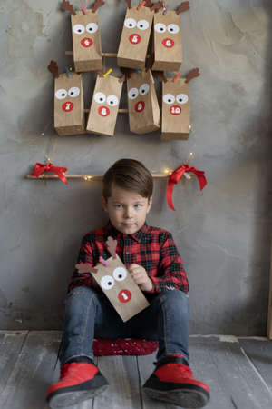 Kids Christmas fun concept. Advent calendar packages hanging on the wall, a child boy sitting on the floor holding a gift in his hands.の写真素材