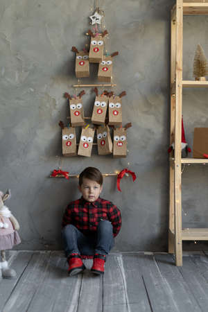 Boy in jeans sits on the floor under the advent calendar hanging on the wall, handmade paper bags with sweets for children, child on eve of christmasの写真素材