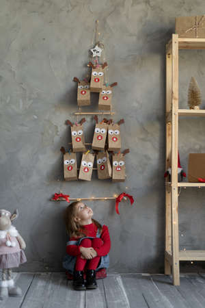 Happy girl looking at advent calendar on the wall sitting on the floor in the room, handmade christmas decor, horizontal view, kid festive fan.の写真素材