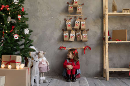 Little girl with a Christmas tree in the living room with an advent calendar hanging on the wall in craft bags, gifts and festive decor on the eve.の写真素材