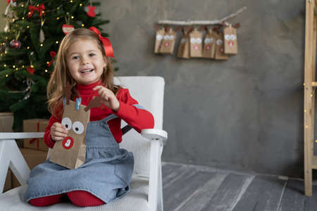 Gift concept for Christmas. Happy little girl with a package of advent calendar sitting at home on an armchair, a Christmas tree near the wall.の写真素材