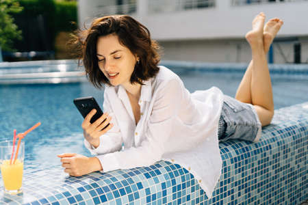 A millennial girl lies with a phone on the edge of the swimming pool in a mans shirt and communicates on the Internet in social networks with friends. Online technologies and communicationsの写真素材