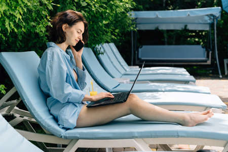 Side view of Caucasian girl working with laptop freelance at resort. Chatting with colleagues sitting by the pool at the hotel. Online saleの写真素材
