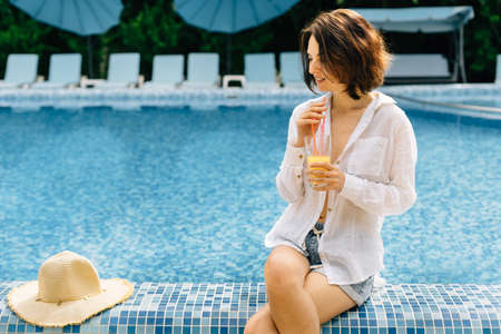 A woman in a shirt drinks freshly squeezed juice from a glass while sitting on the edge of the swimming pool during her summer vacation or relaxing by the water. Latina girl heals with juice.の写真素材
