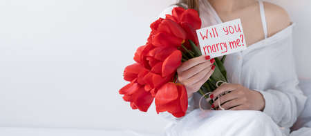 Girl with flowers and a postcard will you marry me on a white background. Bouquet of red tulips in hands. Festive concept Valentines Day. Banner.の写真素材
