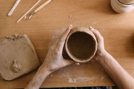 pottery in workshop clay art concept. close-up on the hands of a young ceramist with an unbaked clay pot. artisan hands hold a clay cup and a decoration tool.の写真素材
