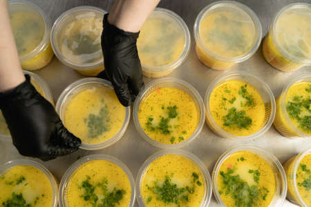 many disposable plates with soup in a lunch box stand on the table in the kitchen in a restaurant female hands are covering the food with a lid. Takeaway food concept during quarantine or pandemic.の写真素材
