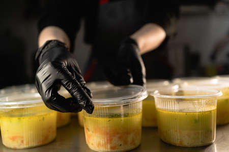 Food delivery. Disposable plates of soup in a lunch box are on the table in the kitchen in a restaurant. Female hands are covering with a plastic lid. Takeaway food conceptの写真素材