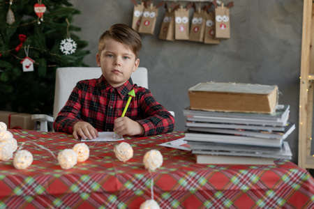 Christmas time, a boy writing a letter to Santa Claus, a child at home with a pen writes a list of New Years gifts, portrait school boy at the tableの写真素材