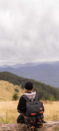 A woman with a backpack sitting on the slope on a fallen tree and enjoying the scenic view. Hiking and climbing to the top of the mountain.の写真素材