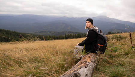 A tourist traveling in the mountains. Green hills and mountains. A girl resting during a trip through the mountains sits on a fallen tree and looks at the beautiful mountain landscape.の写真素材