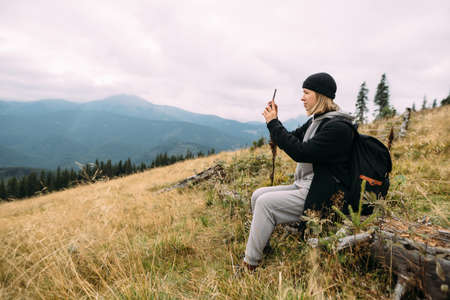 Happy tourist woman sits on top of a mountain during a halt, taking a photo on a mobile smartphone, Blogger, looking at the mountains.の写真素材
