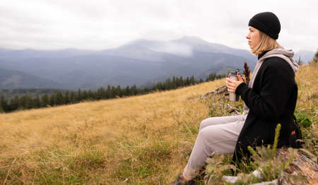 A woman traveler on top of a high mountain drinks coffee from a metal mug, girl looks at the mountains. Free tourist girl, background mountainの写真素材