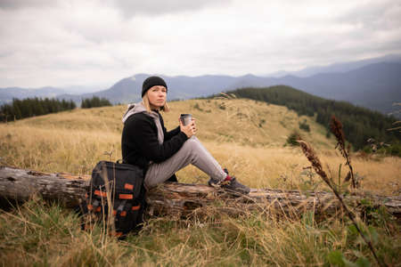Young woman with a thermos cup resting on a fallen tree in the mountains. Freedom, food, travel and vacation concept, outdoor activities.の写真素材
