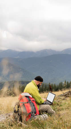 a bearded Caucasian man with a laptop sits on a fallen tree in the mountains with a beautiful view of the valley.の写真素材