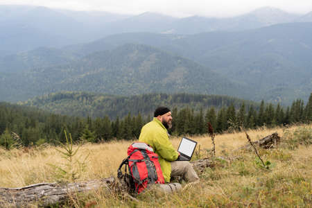 a bearded Caucasian man with a laptop sits on a fallen tree in the mountains with a beautiful view of the valley.の写真素材
