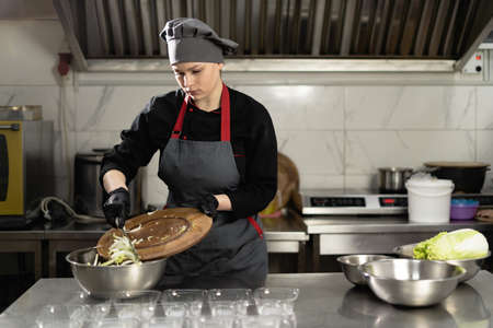 food delivery. A female chef in a restaurant prepares takeaway food in disposable plastic trays. Pour the chopped cabbage into a bowl.の写真素材
