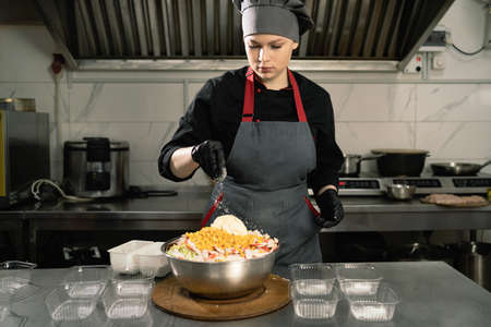 takeaway food in the restaurant. Woman chef in a restaurant in the kitchen pour salt into a large bowl of salad, disposable plastic trays are on the table. Food delivery.の写真素材