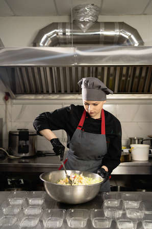 takeaway food in the restaurant. A woman chef in a restaurant is dressed in a uniform in the kitchen, stirring a spoonful of salad in a large bowl, disposable plastic trays are on the table. Food delivery. Copy space.の写真素材