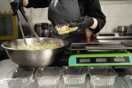 Food delivery in the restaurant. Close-up of female hands in gloves preparing portioned takeaway food. Weigh the salad in a disposable plastic container on an electronic scale.の写真素材