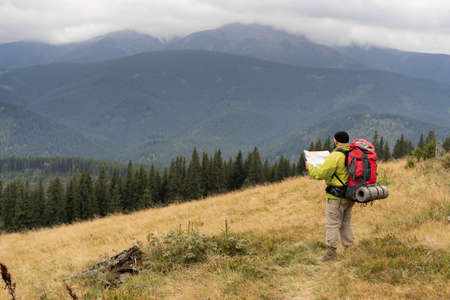 bearded Caucasian male tourist on stands with a map on a mountain trail while hiking in the mountains.の写真素材