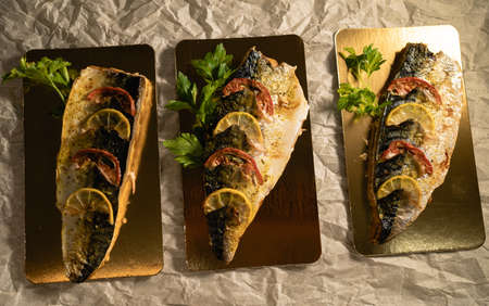 grilled fish mackerel under lemon with herbs and tomatoes, packaging on cardboard for food delivery, sea fish in a restaurant for dinner, appetizing fish filletの写真素材