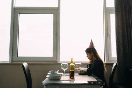 sad lonely birthday girl, lady sitting with cake at home by the window, unhappy birthday girl near the window. Lonely party, remote birthdayの写真素材