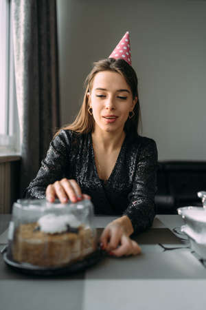 Birthday at home. Young happy woman opens a box with a cake. A girl in a festive cap and dress at the table. Wall background. Birthday celebrationの写真素材