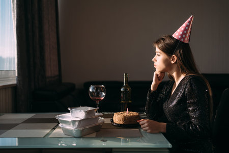 Young woman sitting alone at the table on her birthday. festive hat on the head. The lady feels...