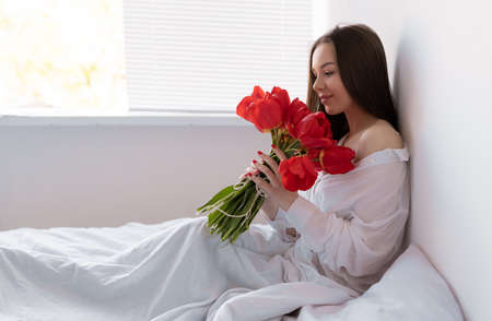 young beautiful woman in the morning in the bedroom sits with a large bouquet of tulips. flowers to a woman for anniversary, valentines day, or just like that.の写真素材