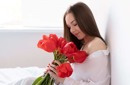 close-up portrait, young beautiful woman in the morning in the bedroom sits with a large bouquet of tulips. flowers to a woman for anniversary, valentines day, or just like that.の写真素材