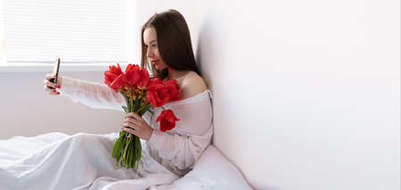Portrait of a sexy girl with a bouquet of red tulips in the morning in the bedroom for Valentines Day makes a selfie on her smartphone. Woman with flowers on the bed Womens day.の写真素材
