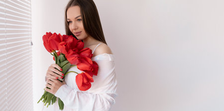 Birthday. Caucasian girl with a bouquet of red flowers in the morning in the bedroom. Tulips for your beloved woman. Flowers delivery home.の写真素材