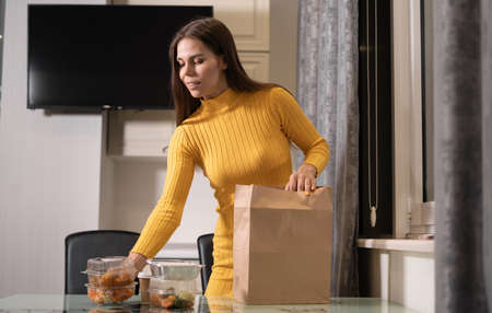 A beautiful Caucasian girl at home in the evening at home takes disposable lunch boxes with food from a paper bag and puts them on the table. Takeaway food concept. Copy space.の写真素材