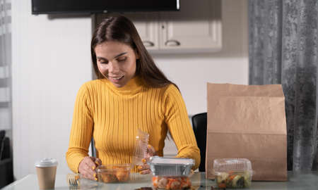A beautiful Caucasian girl is sitting at the table at home alone in the evening and is going to have dinner from disposable dishes. fried chicken legs. The concept of ordering food home. copy space.の写真素材
