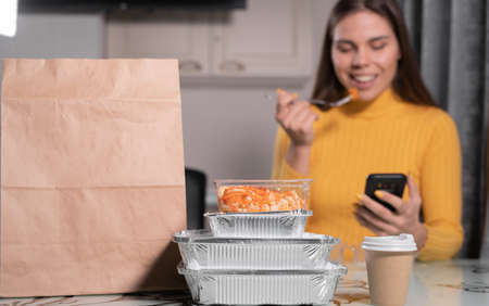 Close up of table with fast food delivery meal and woman holding smartphone while eating. boxes with food in the background. copy spaceの写真素材