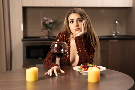 A beautiful woman in a red dress sits at home at a table with a glass of wine, she looks into the camera with a sad look. There are candles on the table nearby. The concept is lonely on valentine's day or anniversary. Copy space.の写真素材