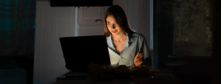 A lonely woman in casual clothes sits at the table at home at night and watches online TV with a smile on her face. Baner. copy space.の写真素材